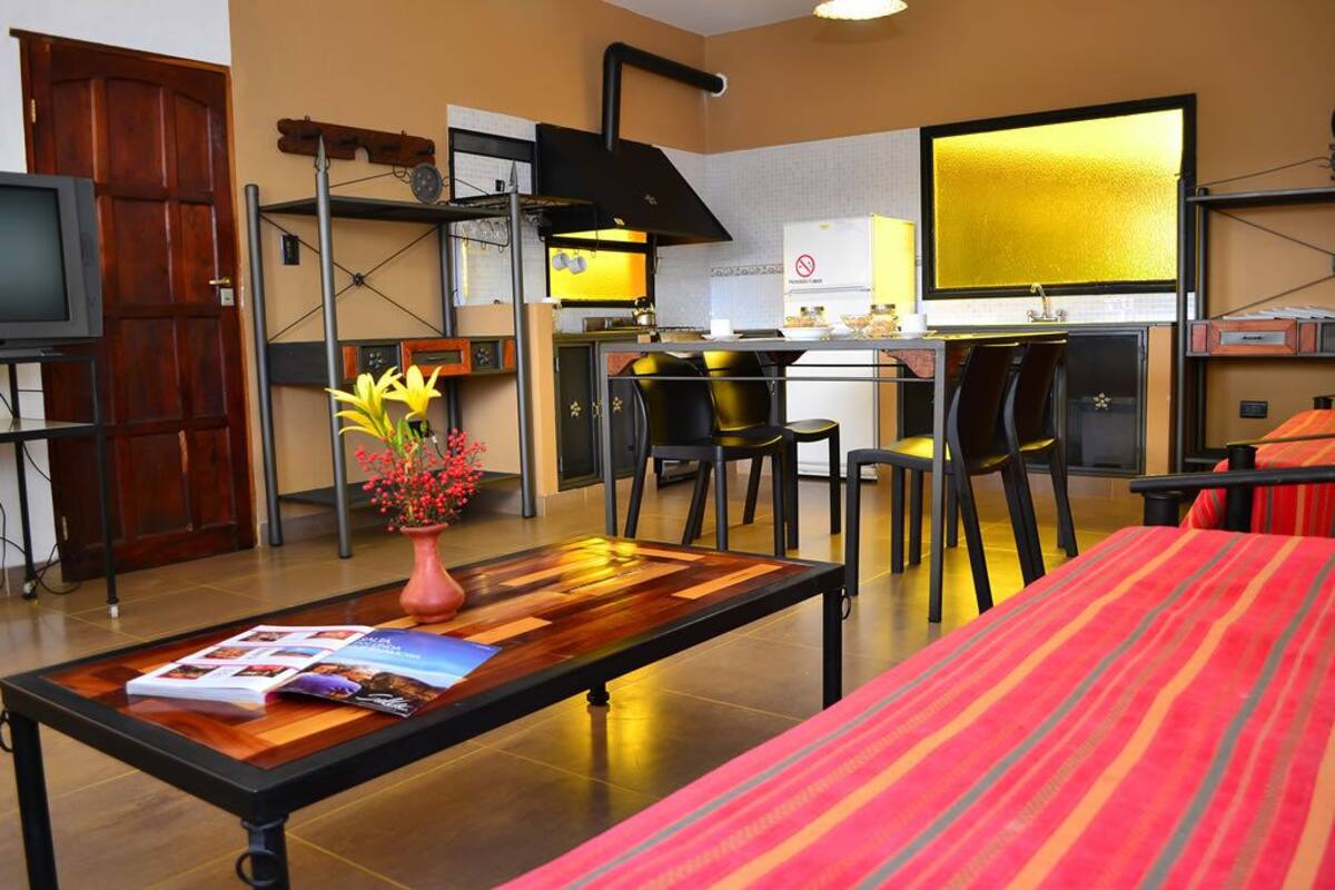 A compact hotel room with a bed, a small dining table, a coffee table, a TV, and a kitchenette.