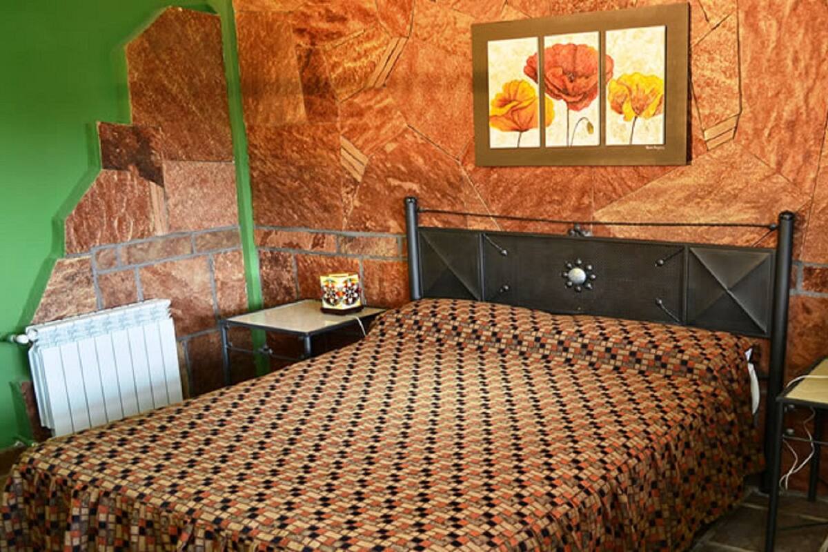 A bedroom with a patterned bedspread, a radiator, a framed picture of flowers, and a small table with a lamp.