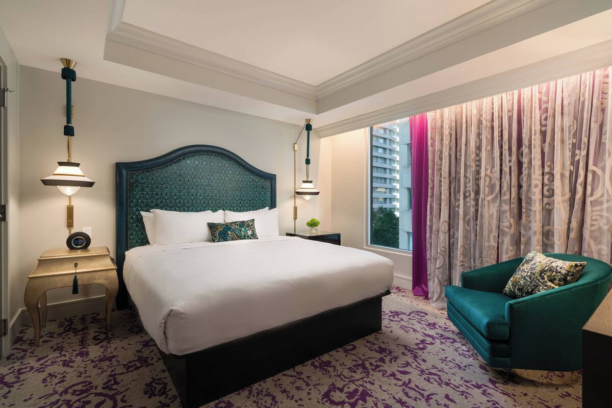 Ein Hotelzimmer mit einem großen Bett, zwei Nachttischen, einem grünen Sessel und einem Fenster mit Vorhängen.