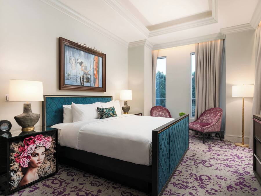 A hotel room with a large bed, two bedside lamps, a painting above the bed, a chair, and a view of the outdoors through a window.