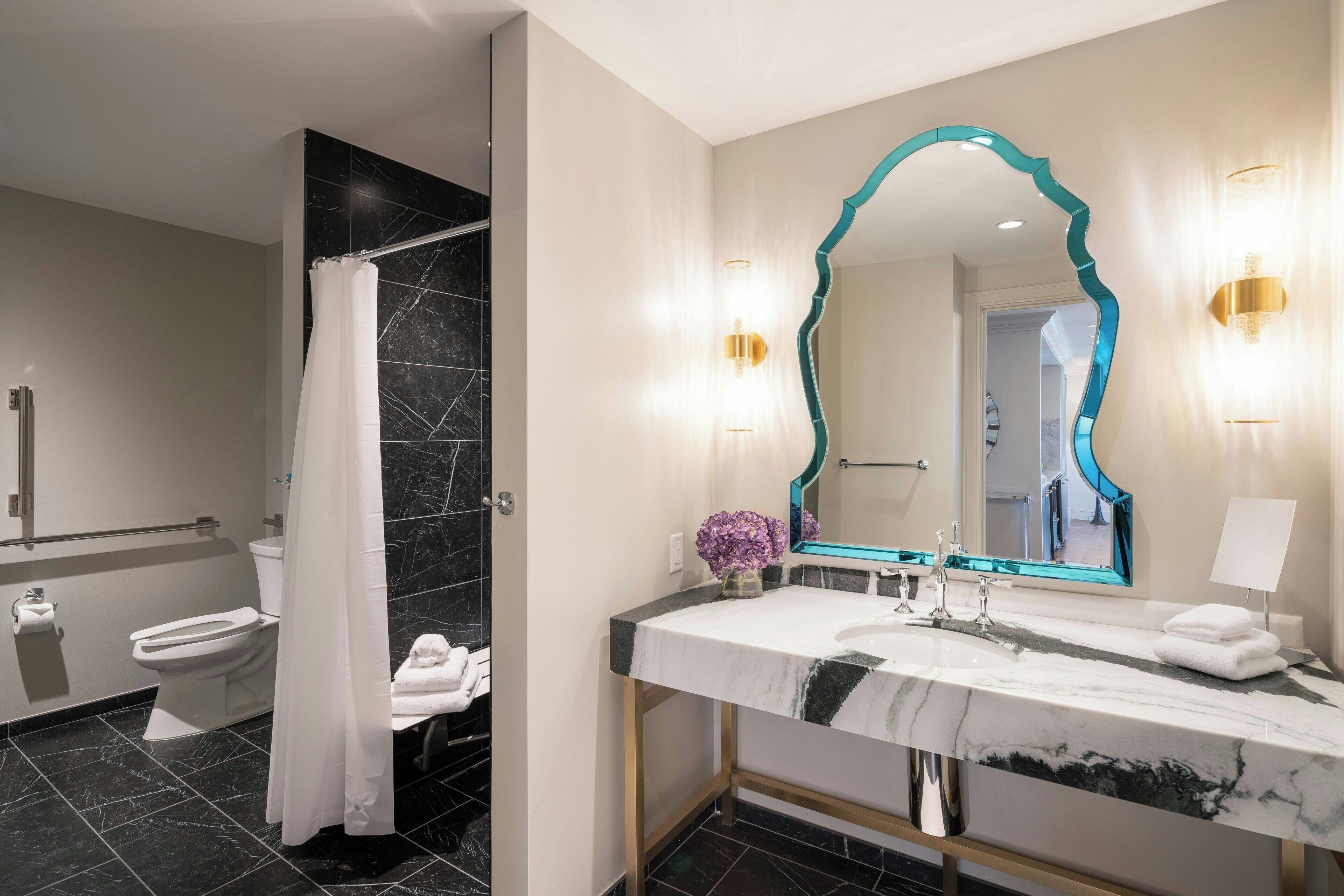 Un baño moderno con lavamanos de mármol, un espejo grande con marco verde azulado y una zona de ducha con azulejos negros.
