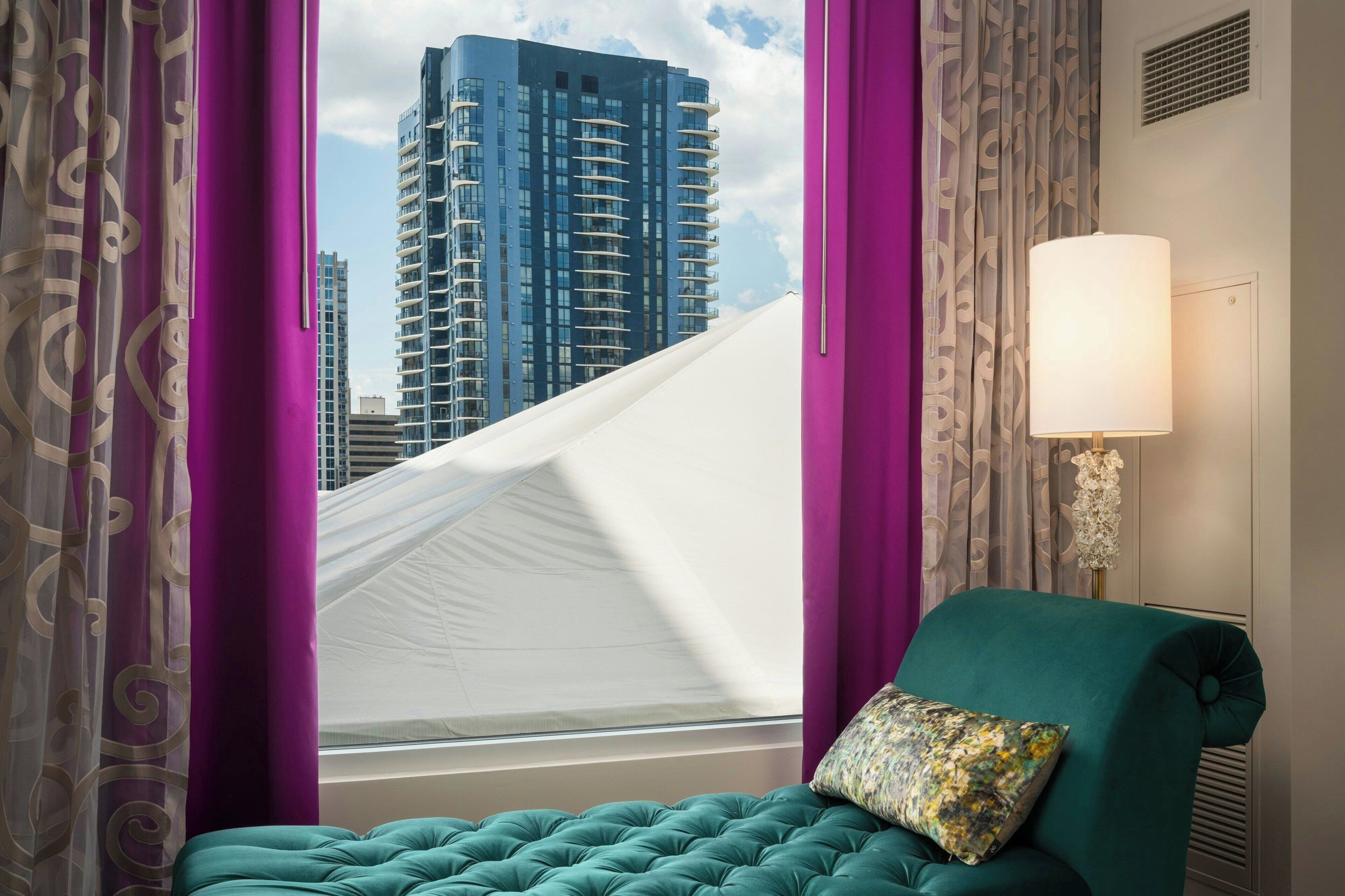 Quarto com cama, cortinas roxas, abajur e vista para um prédio alto.