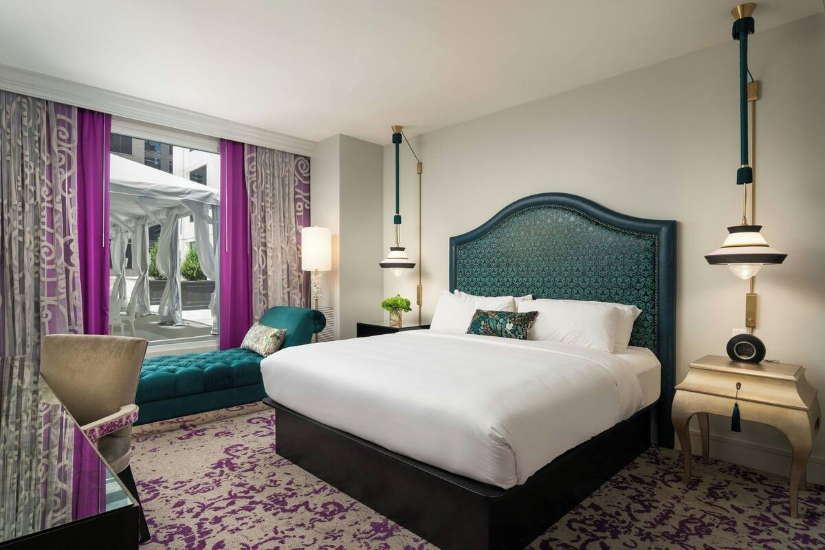 Ein Hotelzimmer mit einem großen Bett, einem blaugrünen Sessel, einem kleinen Tisch mit Lampe und Blick ins Freie durch ein Fenster mit violetten Vorhängen.
