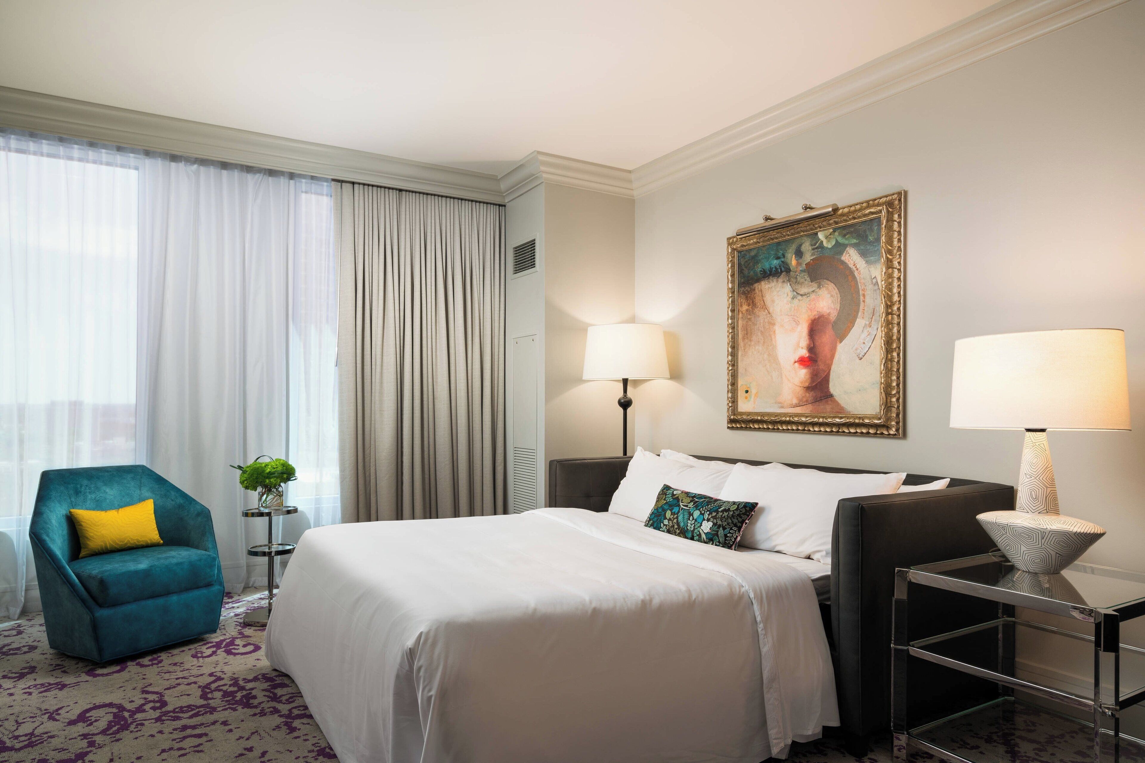A hotel room with a bed, a blue armchair, a side table with a lamp, and a painting on the wall.
