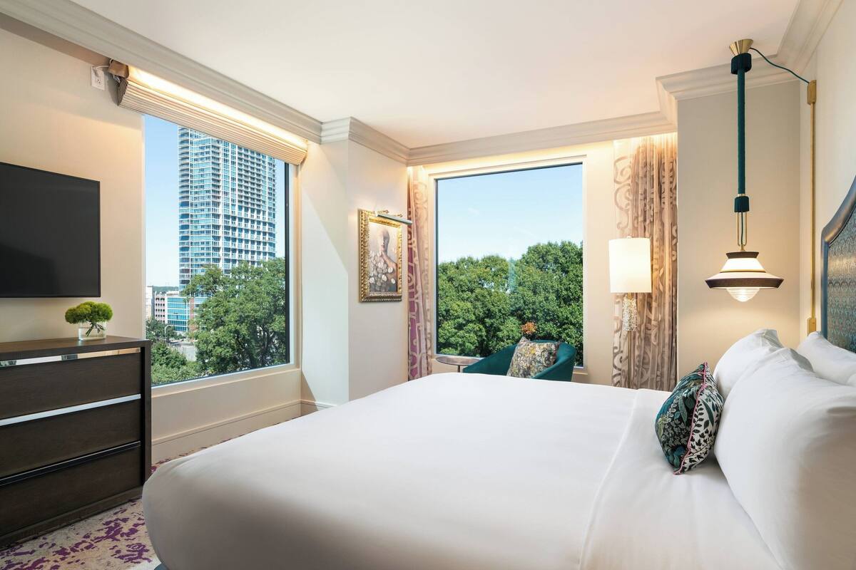 Ein Hotelzimmer mit einem großen Bett, einem Schminktisch, einem Flachbildfernseher, einem Fenster mit Blick auf die Stadt und einer Lampe.