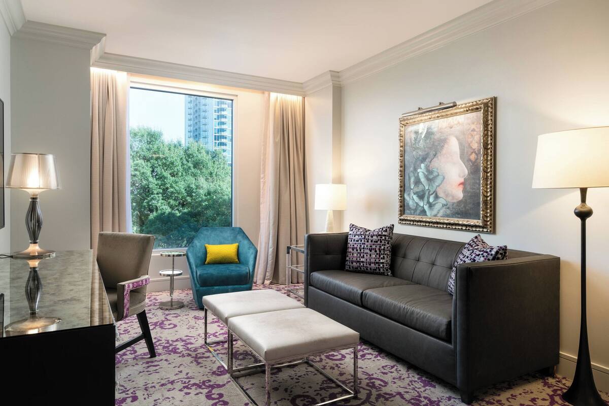 Ein modernes Hotelzimmer mit grauem Sofa, einem blauen Sessel, einem kleinen Tisch und einem großen Fenster mit Blick auf die Stadt.