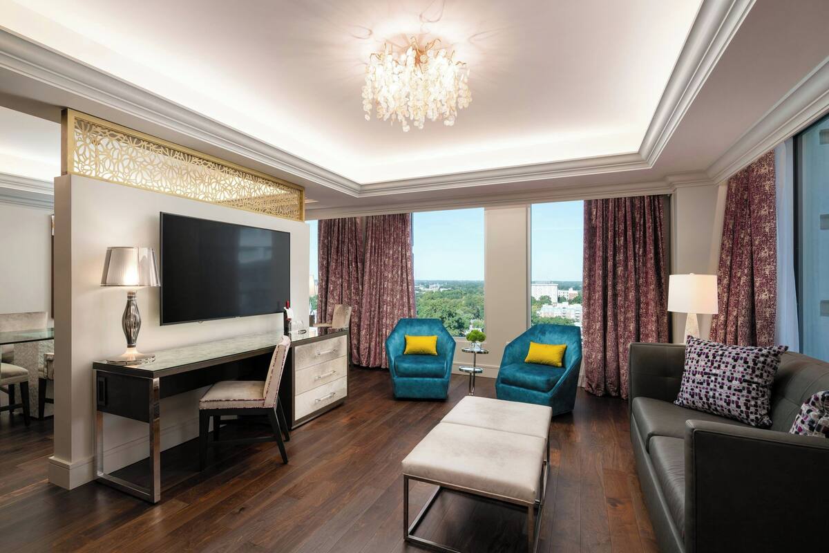 Ein modernes Hotelzimmer mit großem Fernseher, Schreibtisch, Sofa und zwei Sesseln.