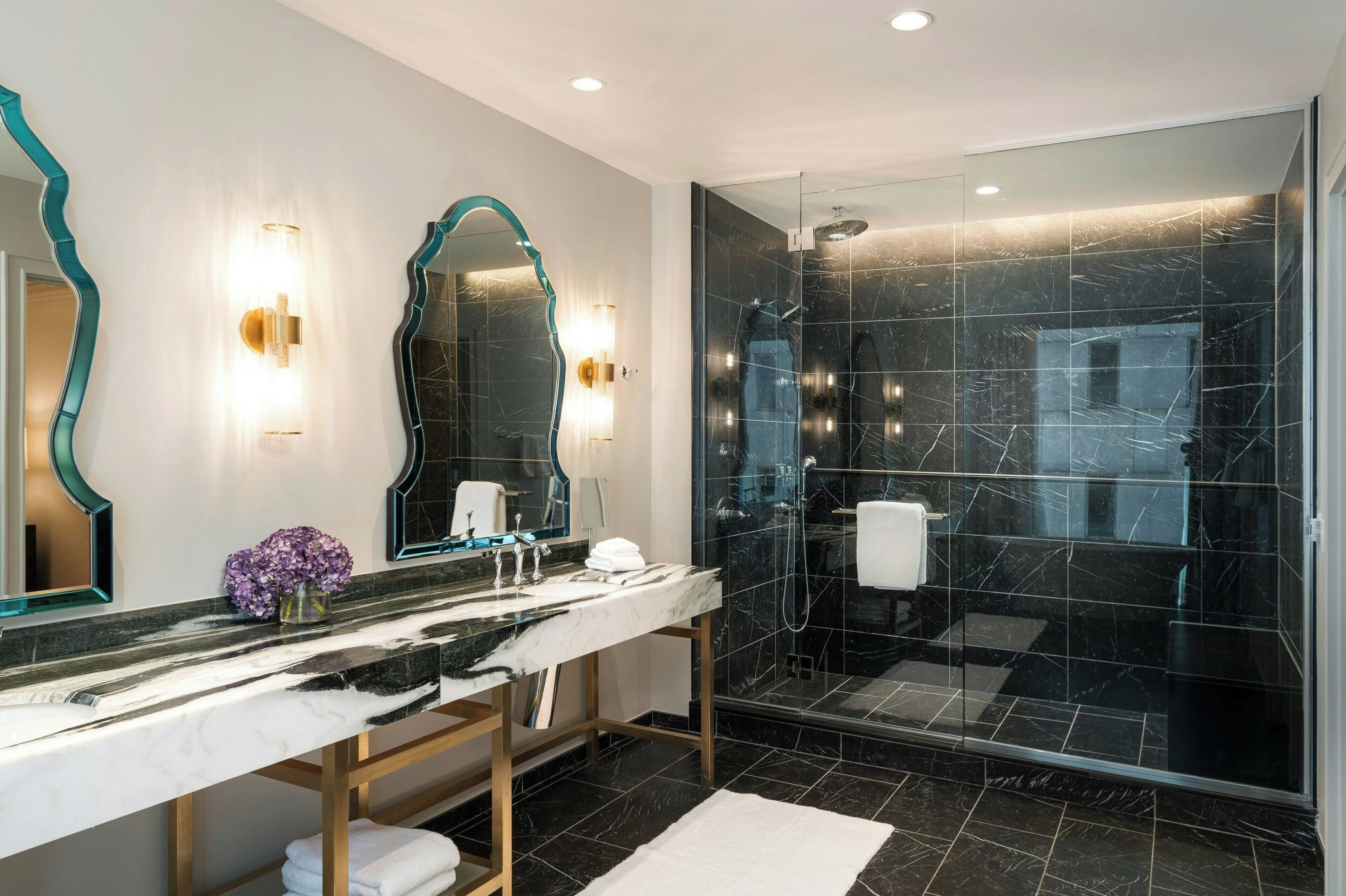 A modern bathroom with a marble vanity, two wall-mounted mirrors, a glass-enclosed shower, and a white bath mat.