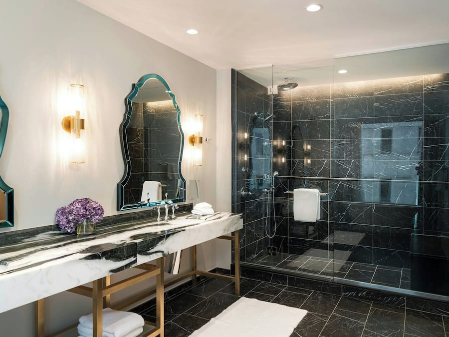 A modern bathroom with a marble vanity, two wall-mounted mirrors, a glass-enclosed shower, and a white bath mat.