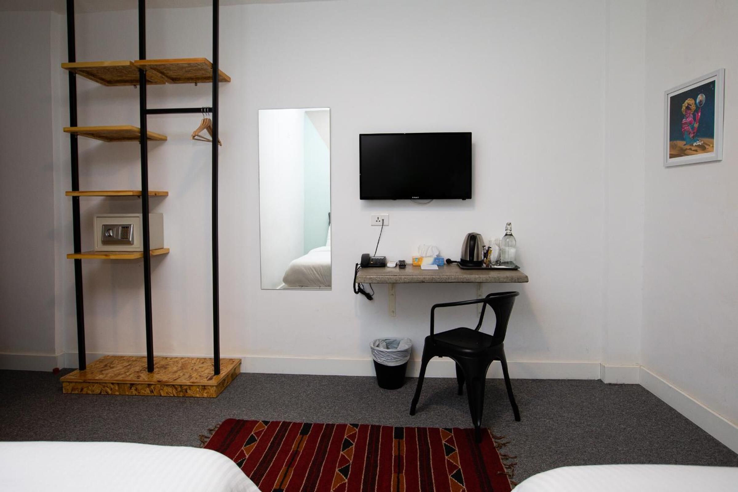A hotel room with a bed, a desk with a chair, a TV, a mirror, and a small trash bin.