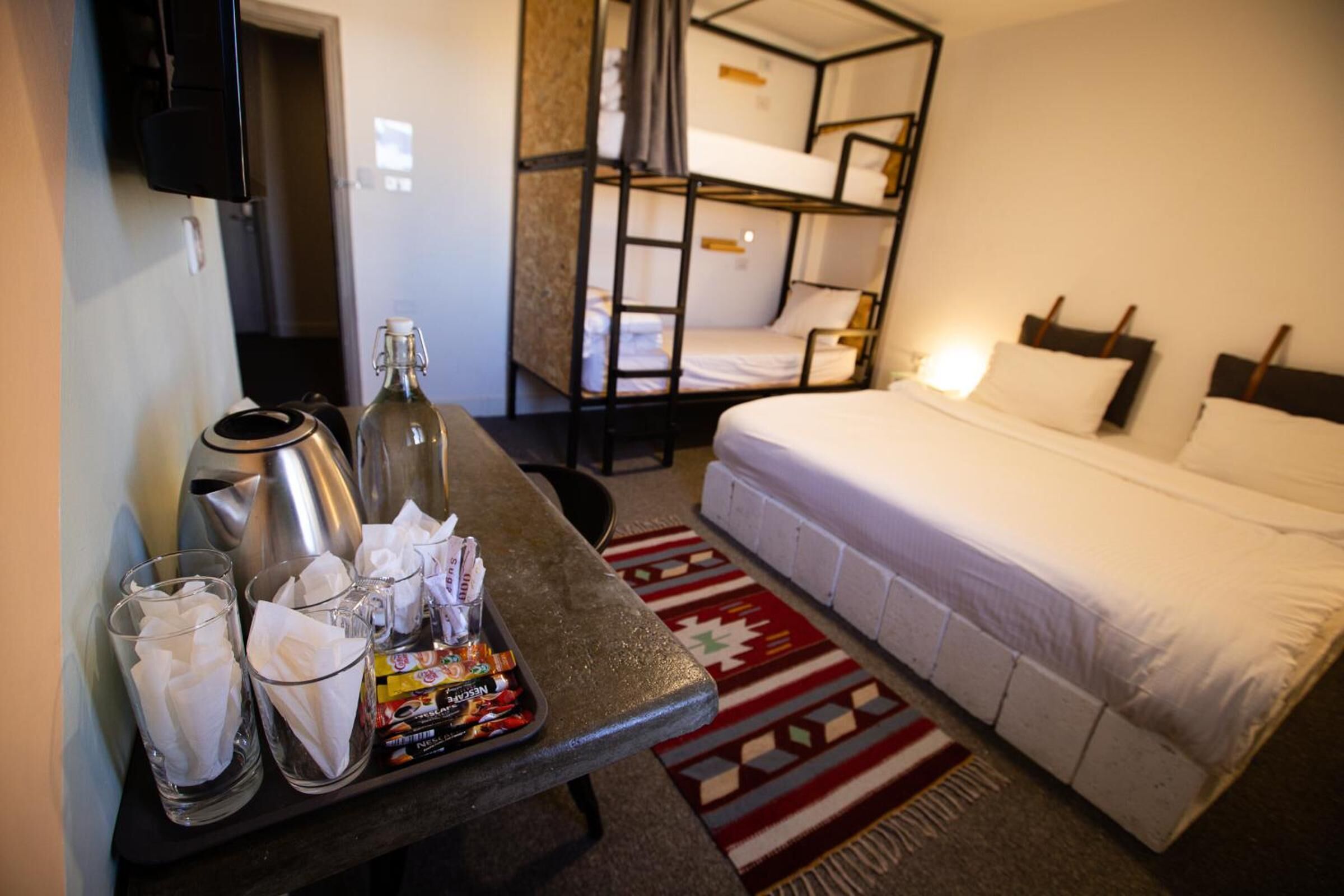Quarto de hotel com beliche, uma cama de solteiro, mesa com bandeja de lanches, uma garrafa e copos.