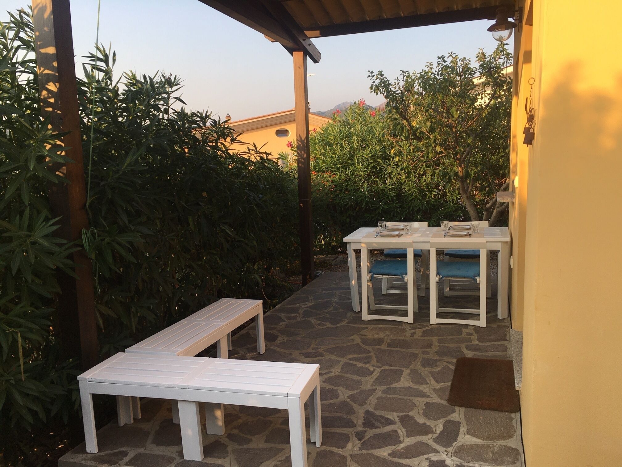Un patio al aire libre con suelo de piedra, muebles blancos y abundante vegetación.