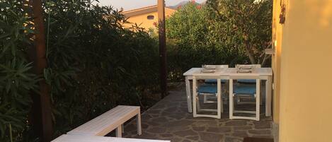 An outdoor patio with a stone floor, white furniture, and lush greenery.