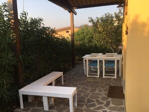 An outdoor patio with a stone floor, white furniture, and lush greenery.