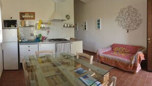 Una pequeña cocina con armarios blancos, un refrigerador y una mesa de comedor con sillas. Hay un sofá en la sala y una puerta de madera.
