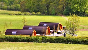 A row of modern, dark-colored cabins with arched openings, set in a lush green landscape with a path leading to a central area with seating and a lamp post.