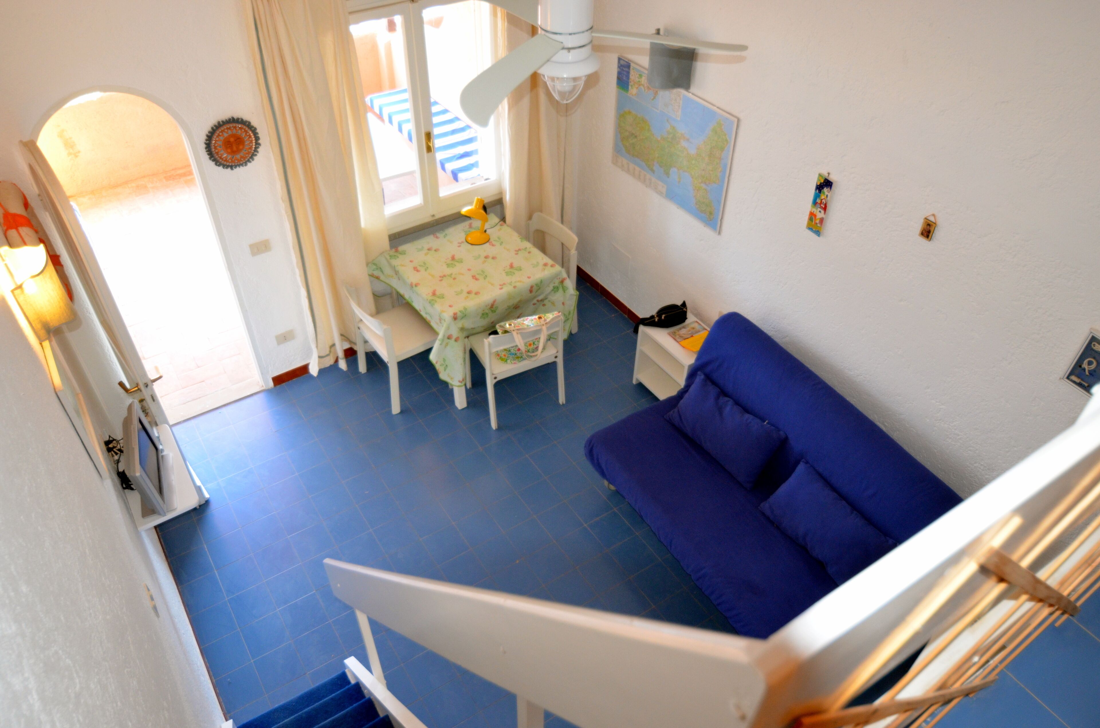 A room with a blue floor, a white sofa, a small table with a lamp, and a ceiling fan.