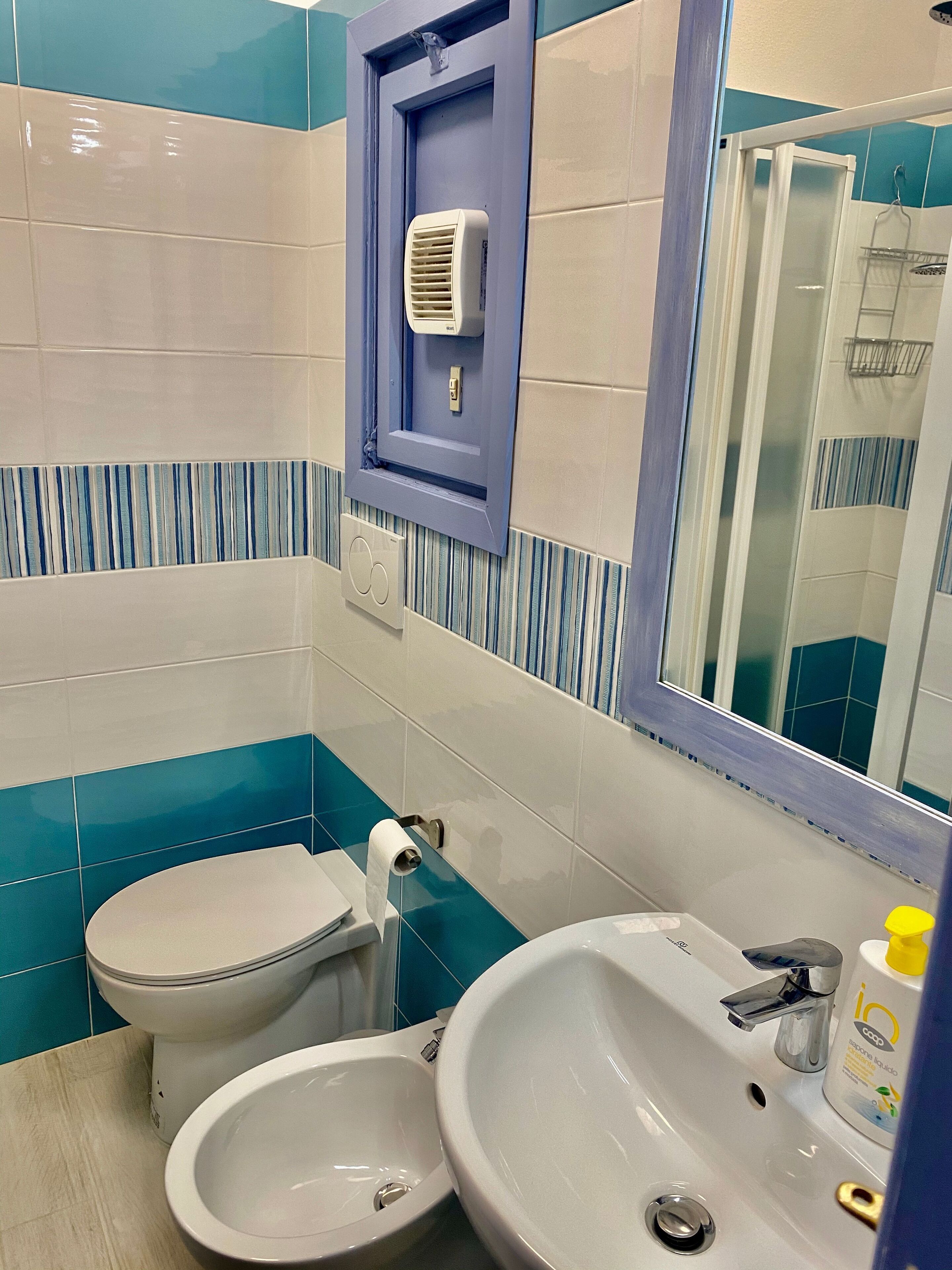 A bathroom with a toilet, bidet, and sink, featuring blue and white tiles, a mirror, and a shower area.