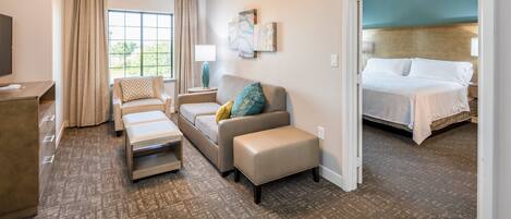 A hotel room with a sofa, ottoman, and a bed. There is a window with curtains, a lamp, and artwork on the wall.
