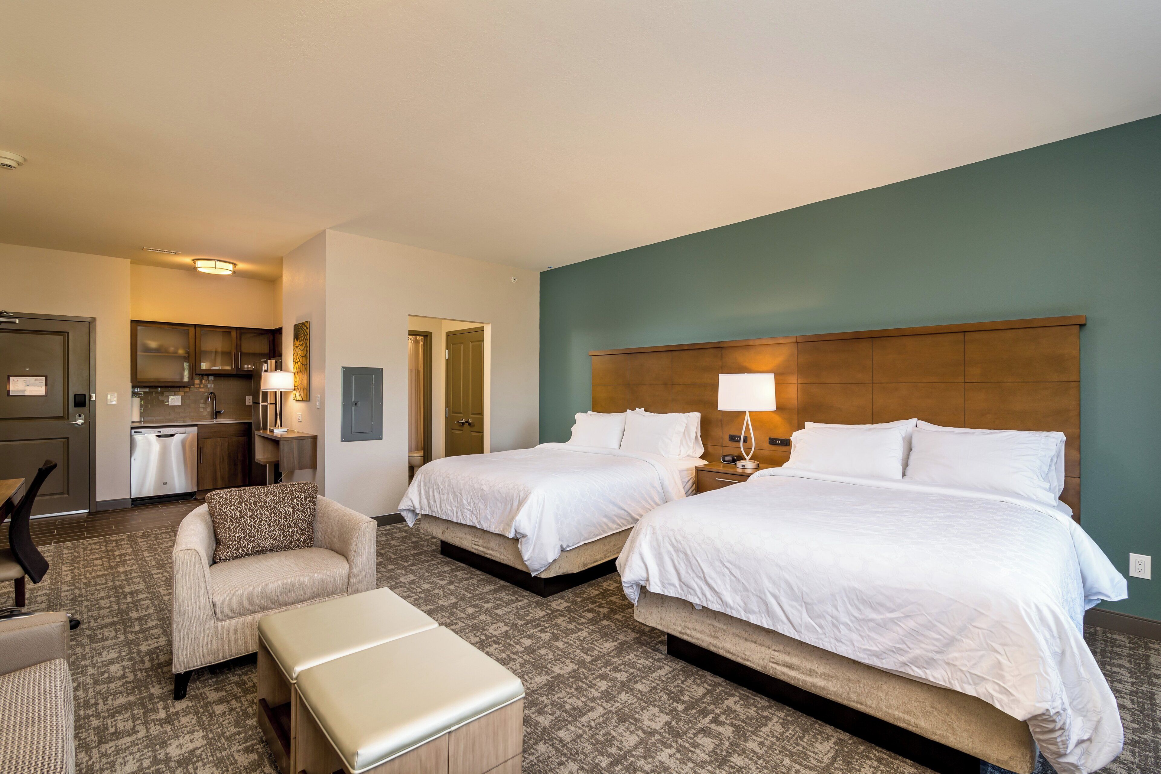 A hotel room with two beds, a kitchenette, a seating area with a chair and ottoman, and a bathroom door.