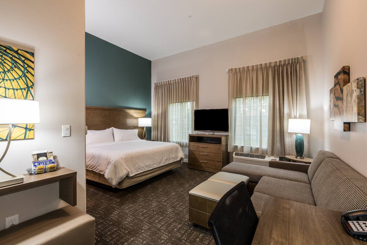 A hotel room with a bed, a sofa, a desk with a lamp, a TV, and a window with curtains.
