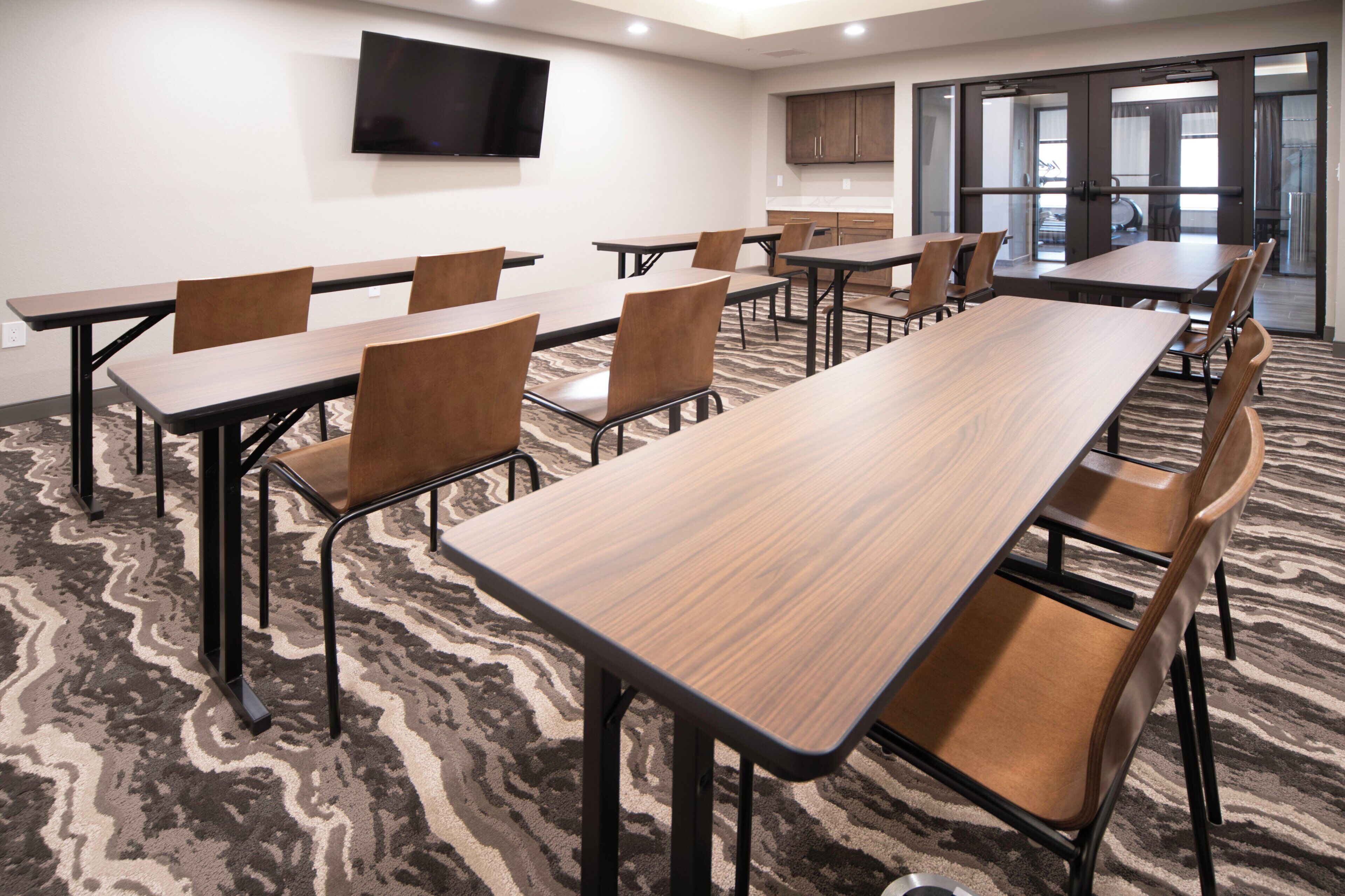 A conference room with multiple tables and chairs, a flat-screen TV mounted on the wall, and a patterned carpet.