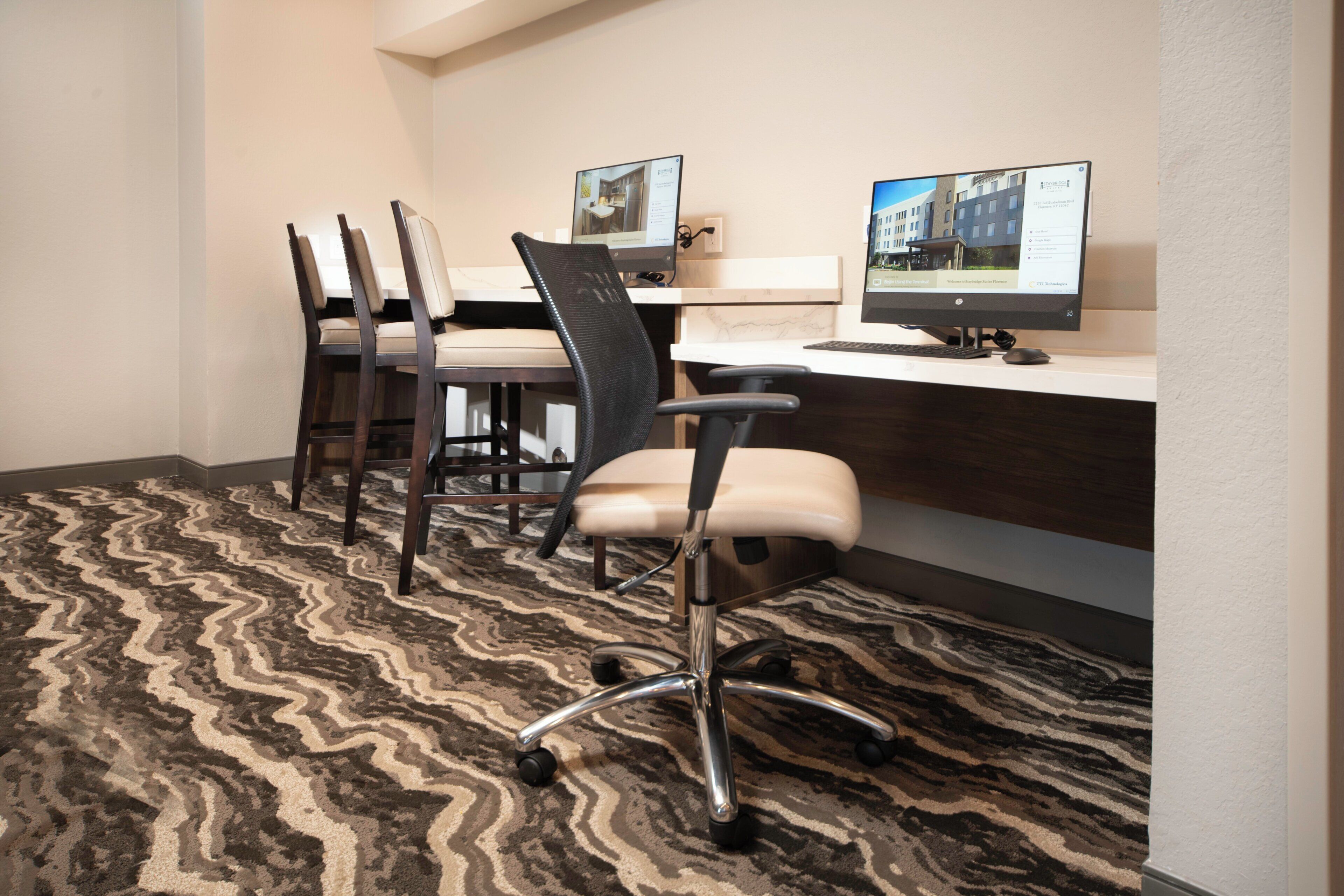 A hotel room with a desk, two chairs, a computer, and a patterned carpet.