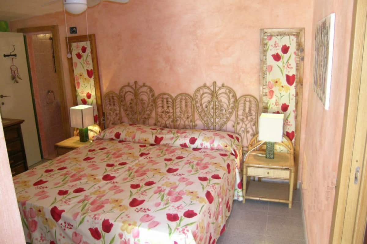 Un dormitorio con colcha estampada, cabecera decorativa, dos mesitas de noche con lámparas y un ventilador de techo.