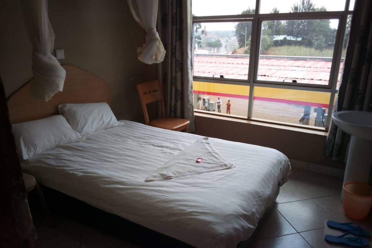 Una cama con sábanas blancas, una silla de madera, una ventana con vista a un edificio de techos rojos y un piso de baldosas.