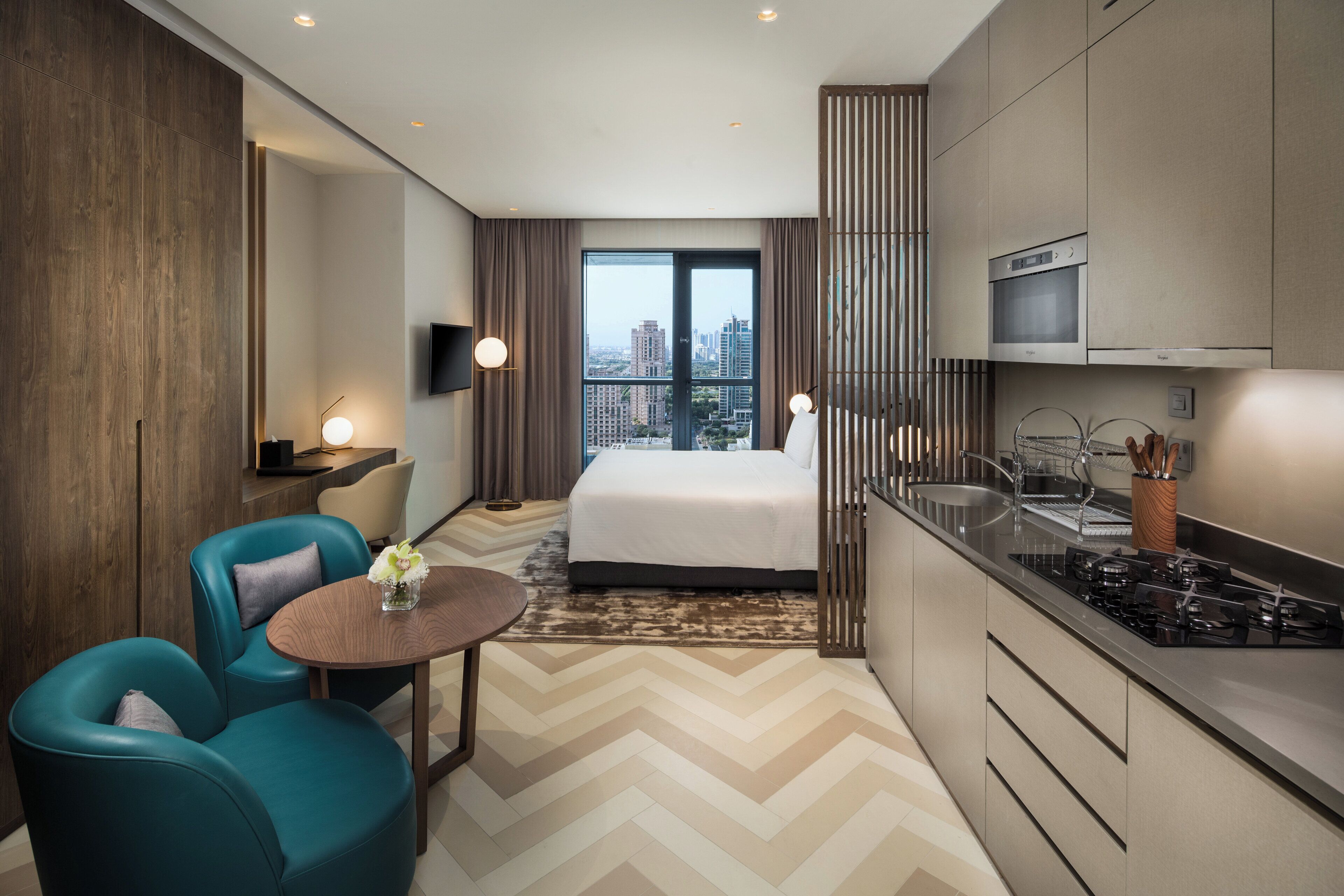Une chambre d’hôtel moderne comprenant un lit, un coin repas avec une table et des chaises, une cuisine équipée d’une cuisinière et une grande fenêtre donnant sur la ville.