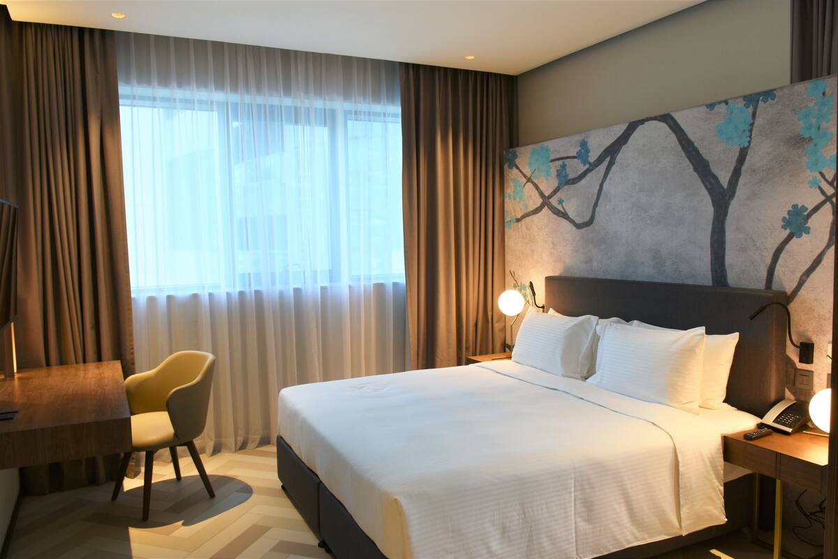 A hotel room with a large bed, a desk with a chair, a bedside table with a lamp, and a painting on the wall.