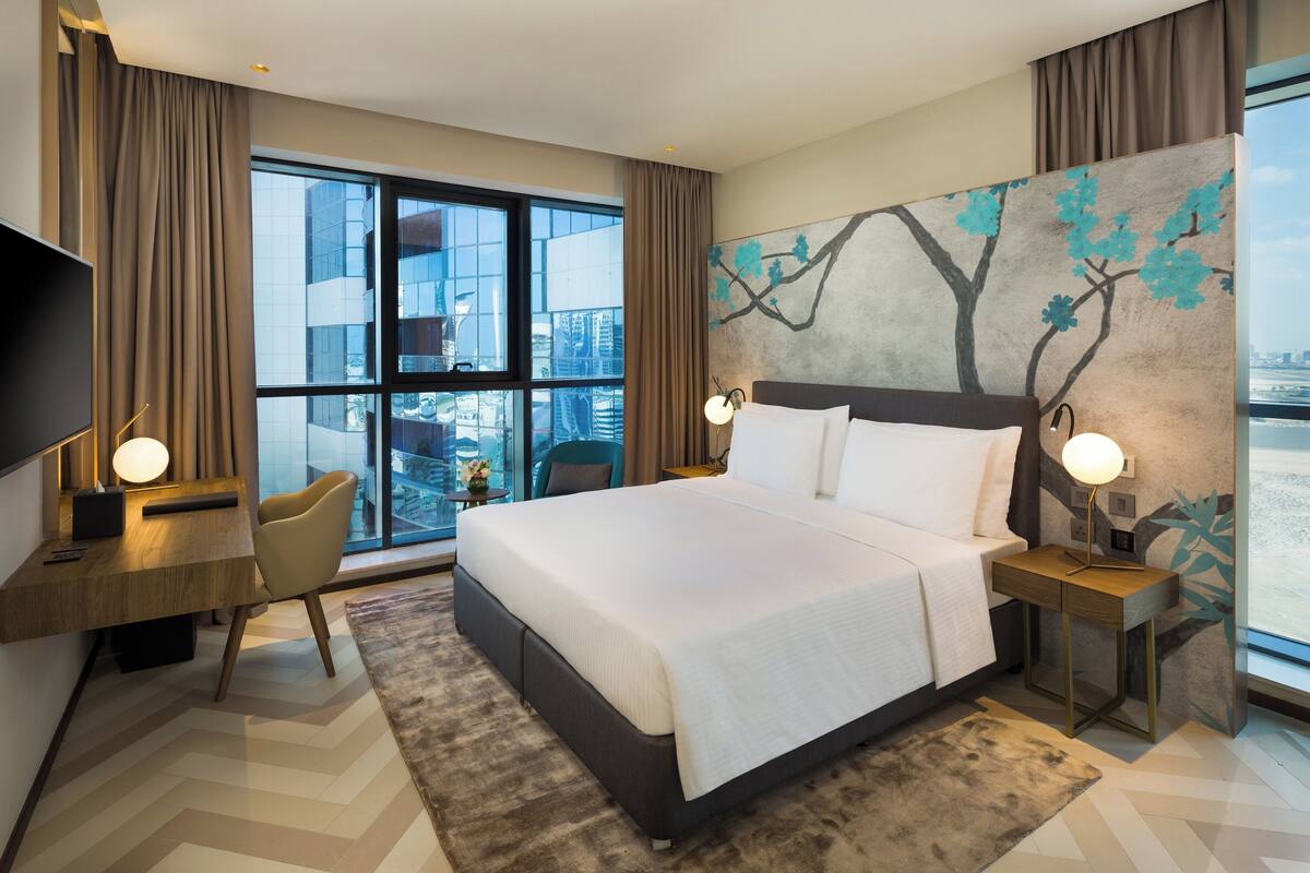 A modern hotel room with a large bed, a desk, and a chair. There is a large window with a view of buildings.