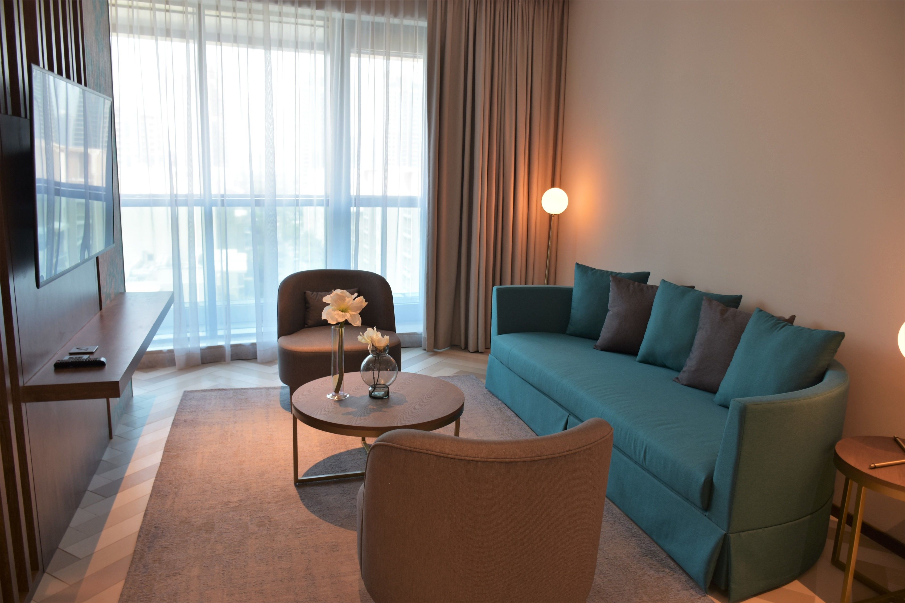 Un salon moderne avec un canapé turquoise, un fauteuil marron, une table basse ronde ornée d’un vase de fleurs et un téléviseur posé sur un meuble.