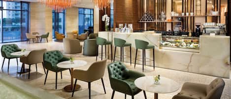 Un moderno hall de hotel con sillones tapizados en tonos verde y beige, un mostrador de recepción de mármol y una barra de café.