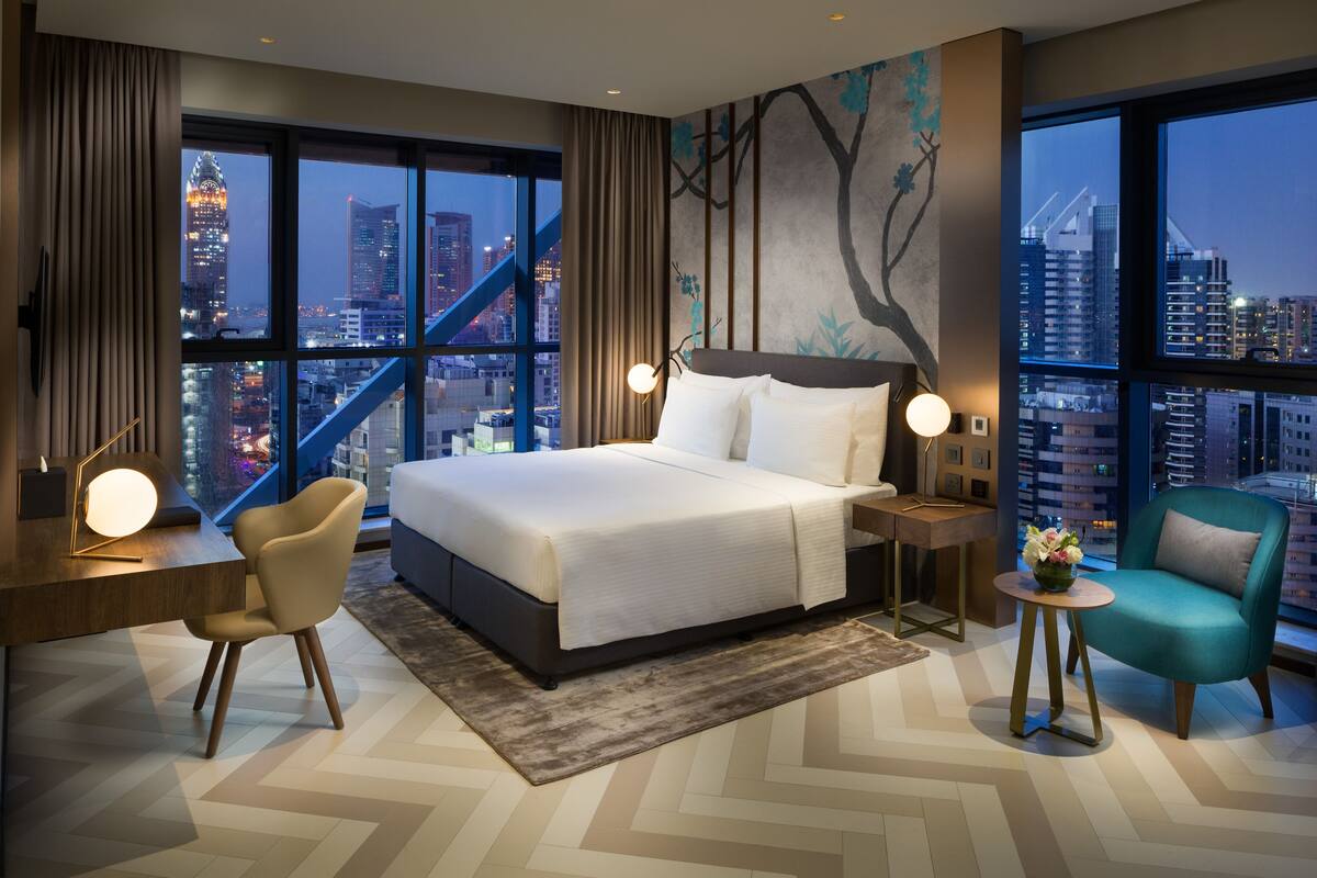 A modern hotel room with a large bed, a desk, a chair, and a view of the cityscape.