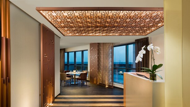 Een moderne hotelgang met een houten vloer, een eetgedeelte met stoelen en een tafel, en een stadszicht door grote ramen.