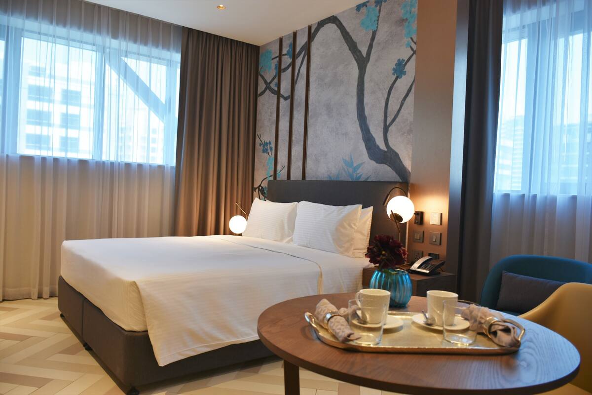 A hotel room with a large bed, a bedside table with a lamp, a small table with a tray of breakfast items, and a wall with a tree mural.