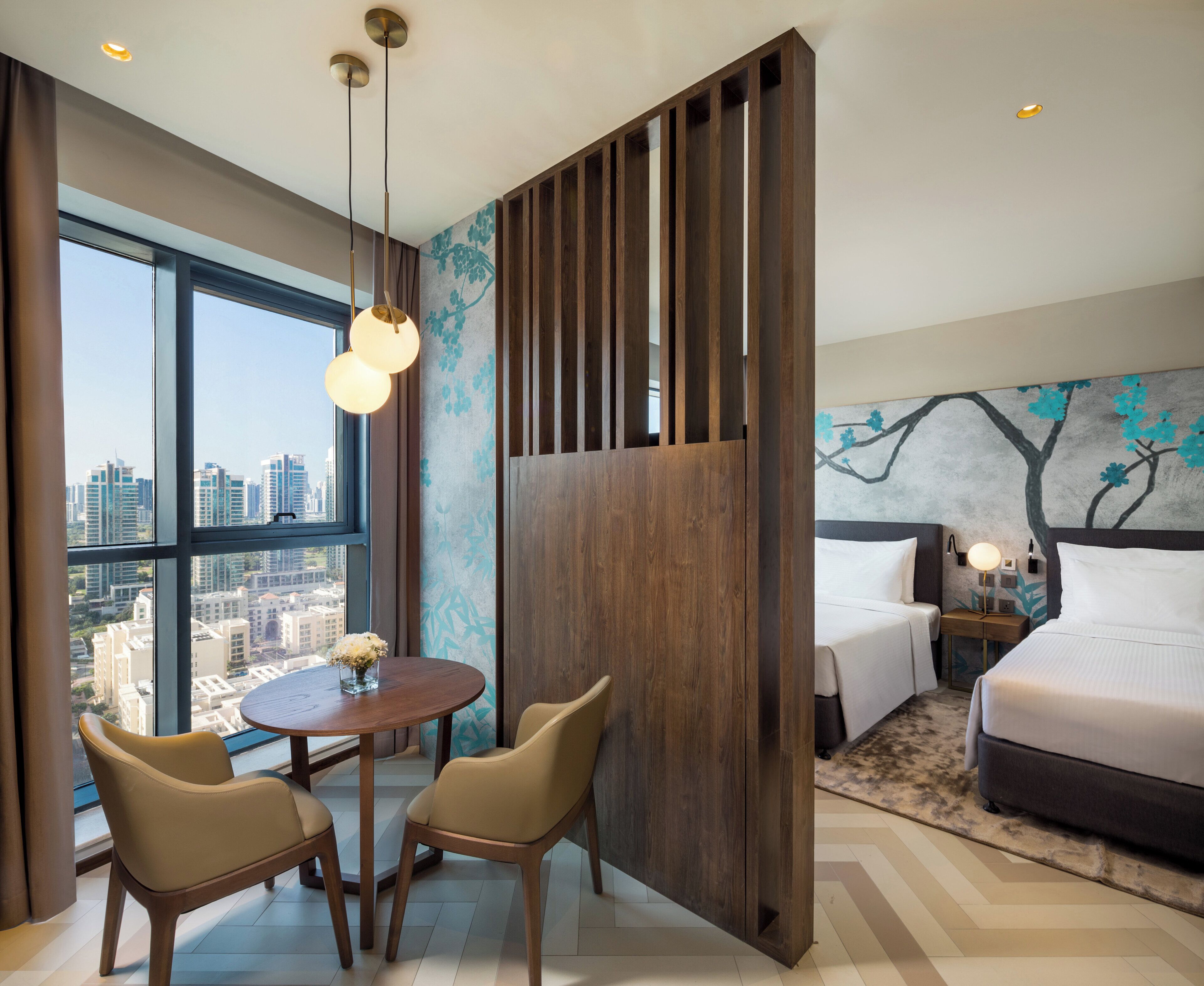 Habitación de hotel con un ventanal grande, una cama, una mesita de noche con lámpara, una mesa redonda de madera con dos sillas y un panel decorativo en la pared.