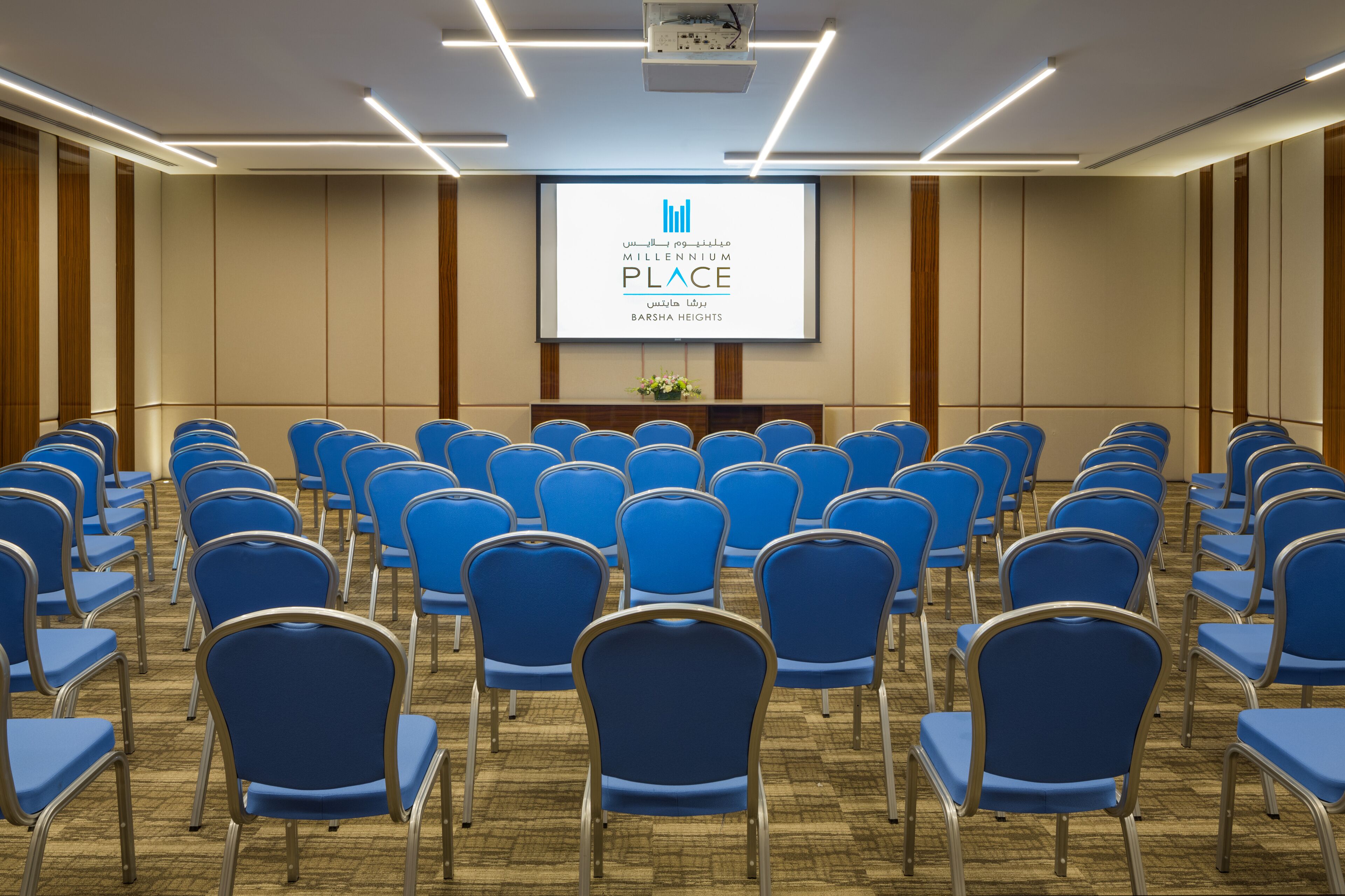 A conference room with rows of blue chairs, a podium, and a screen displaying 'Millennium Place Barsha Heights'.
