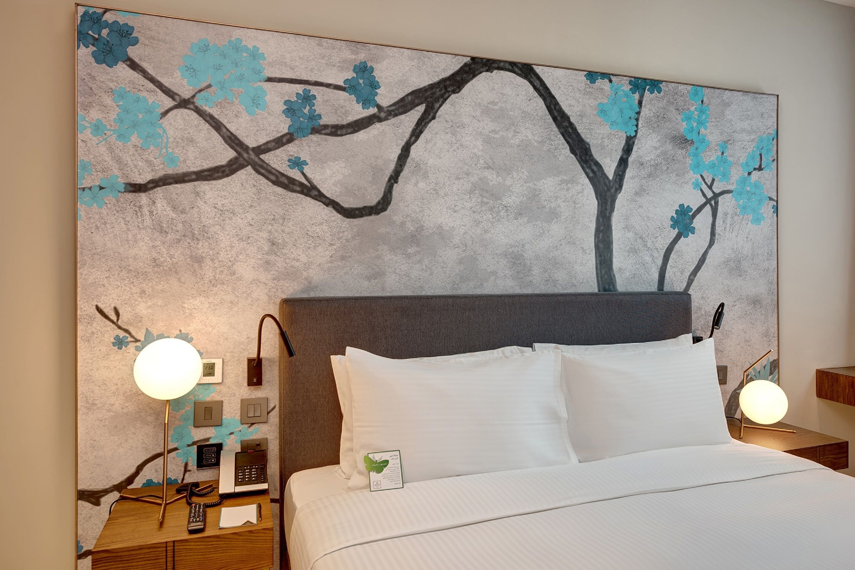 A hotel room with a large painting of a tree with blue flowers on the wall, a bed with white linens, and bedside tables with lamps and a phone.