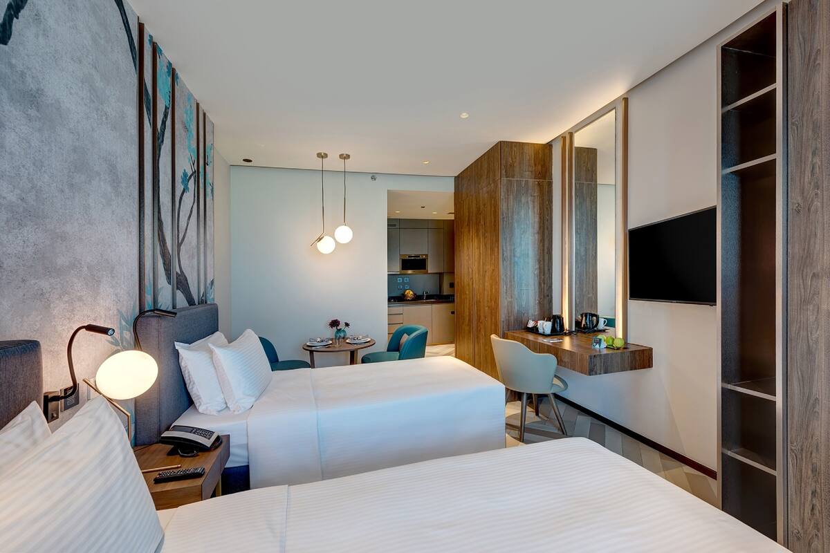 A hotel room with two beds, a desk, a chair, a dining table, a TV, and a kitchen area.