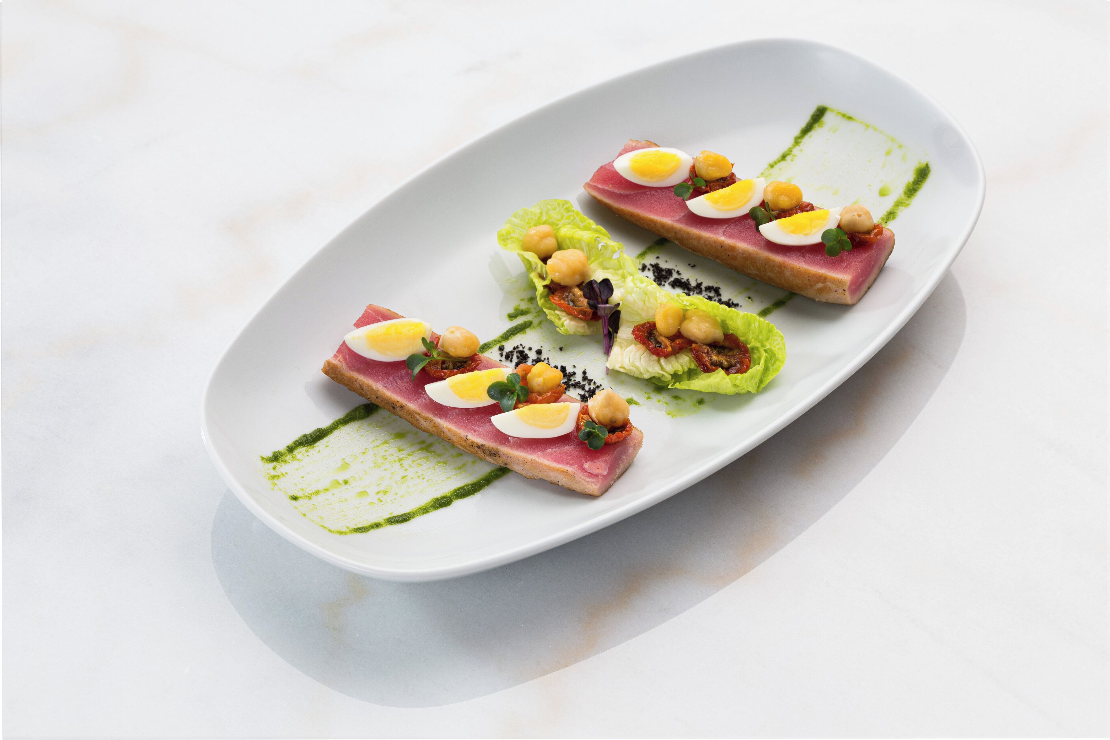 A white oval plate with three pieces of food, each with a slice of pink meat, a boiled egg, and chickpeas, garnished with black seeds.
