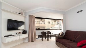 Una sala de estar con televisión de pantalla plana, un sofá, una mesa de comedor con sillas y una ventana con cortinas.