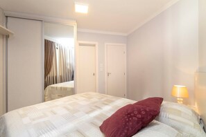 A bedroom with a bed, a mirror, two doors, and a nightstand with a lamp. - Aluguel Apartamento 2 quartos 511 (Itapema)