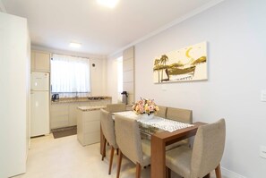 A compact kitchen with a refrigerator, stove, and sink. A dining table with chairs is in the foreground, and a painting is on the wall. - Aluguel Apartamento 2 quartos 511 (Itapema)