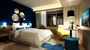 A modern hotel room with a large bed, bedside tables, a wardrobe, a desk with a chair, a small round table with a plant, and a large window with curtains. - Aspira One Sutthisan (Bangkok)