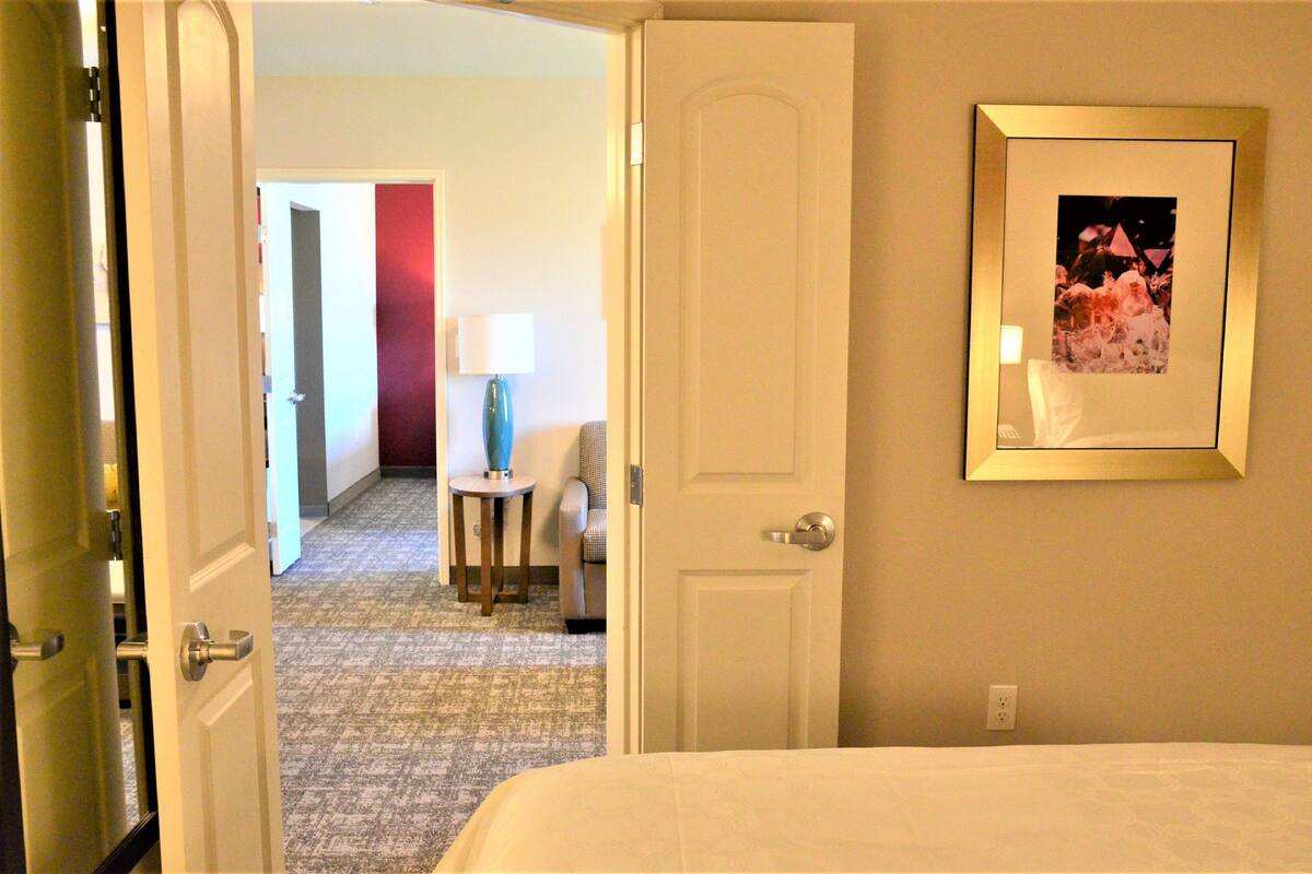 Ein Hotelzimmer mit Bett, Tür, einem gerahmten Bild an der Wand, Lampe und Stuhl.