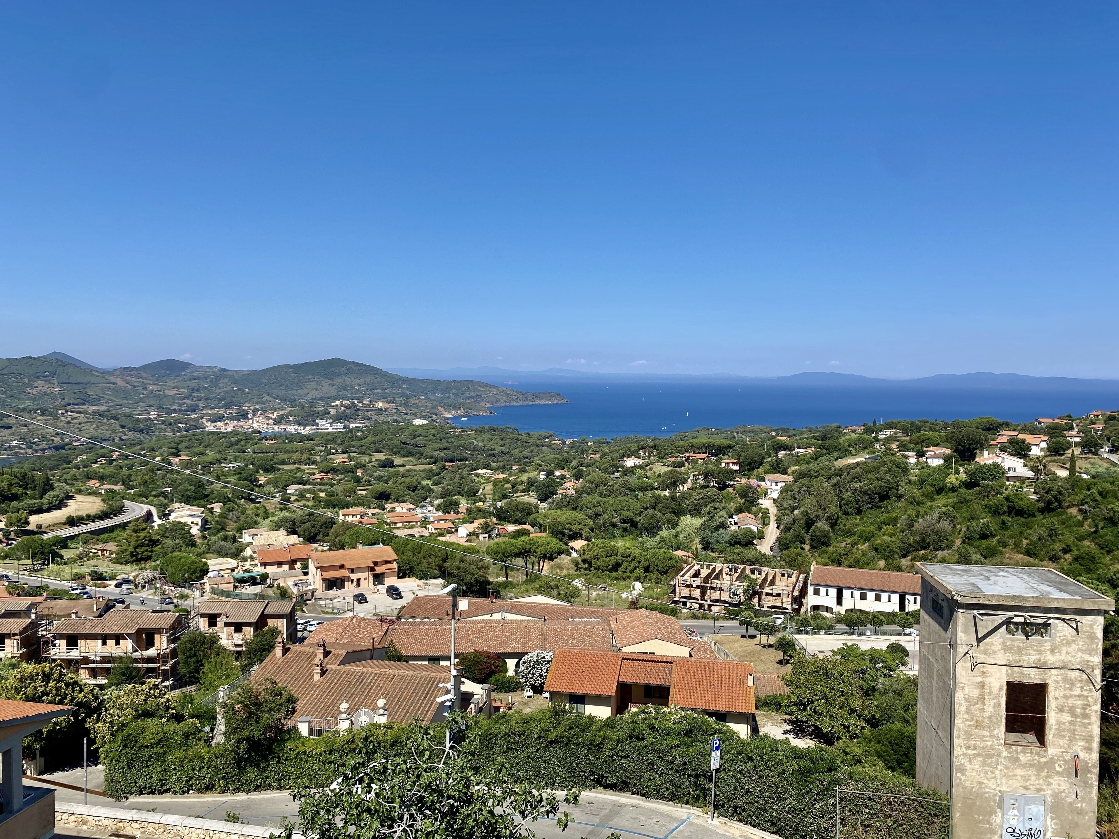 A coastal village with terracotta-roofed houses, a clear blue sea, and distant mountains.
