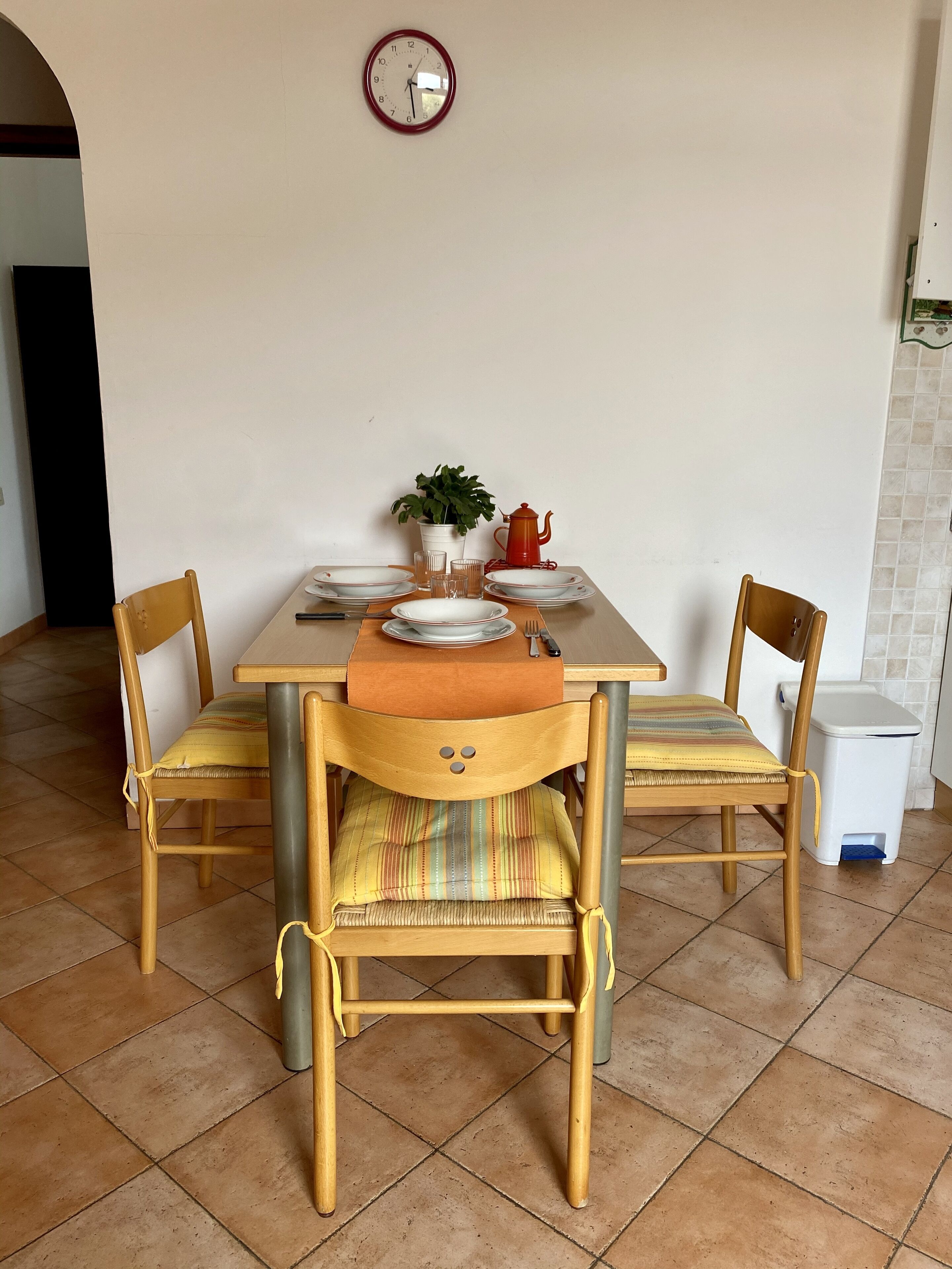 Comedor con mesa dispuesta para cuatro, sillas, un reloj, una planta en maceta y un cubo de basura.