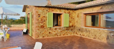 A stone building with green shutters, a tiled patio, and outdoor furniture.